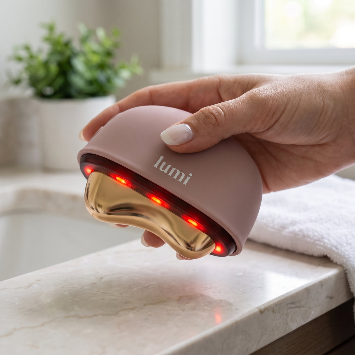 Hand holding the Lumi Ember rosé heated gua sha over a marble bathroom vanity