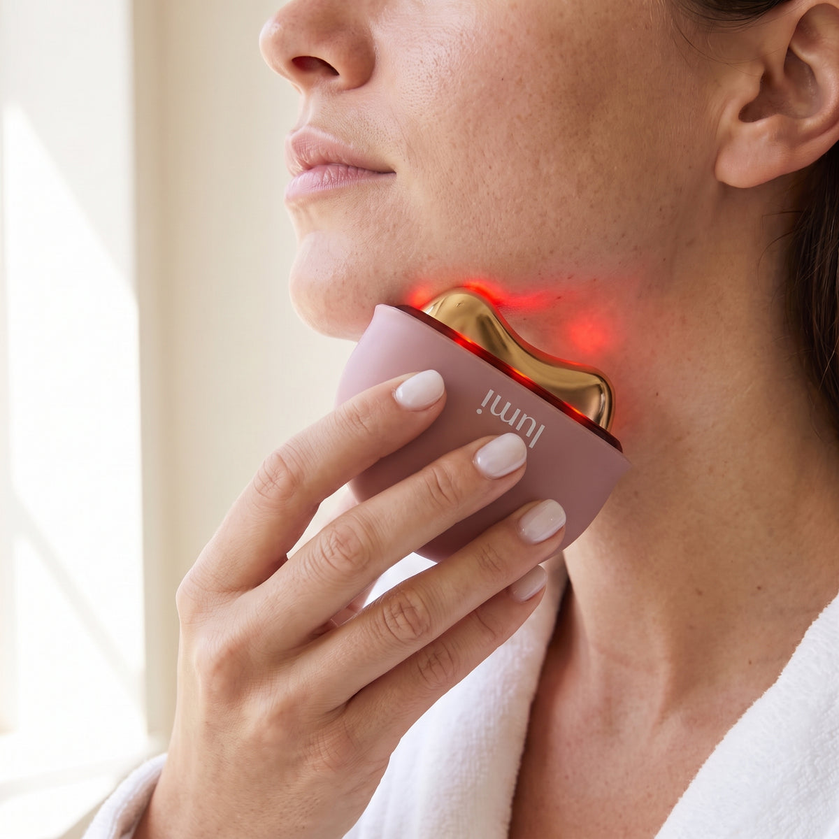 Woman gliding the Lumi Ember heated gua sha along her jawline and neck with a soft red LED glow