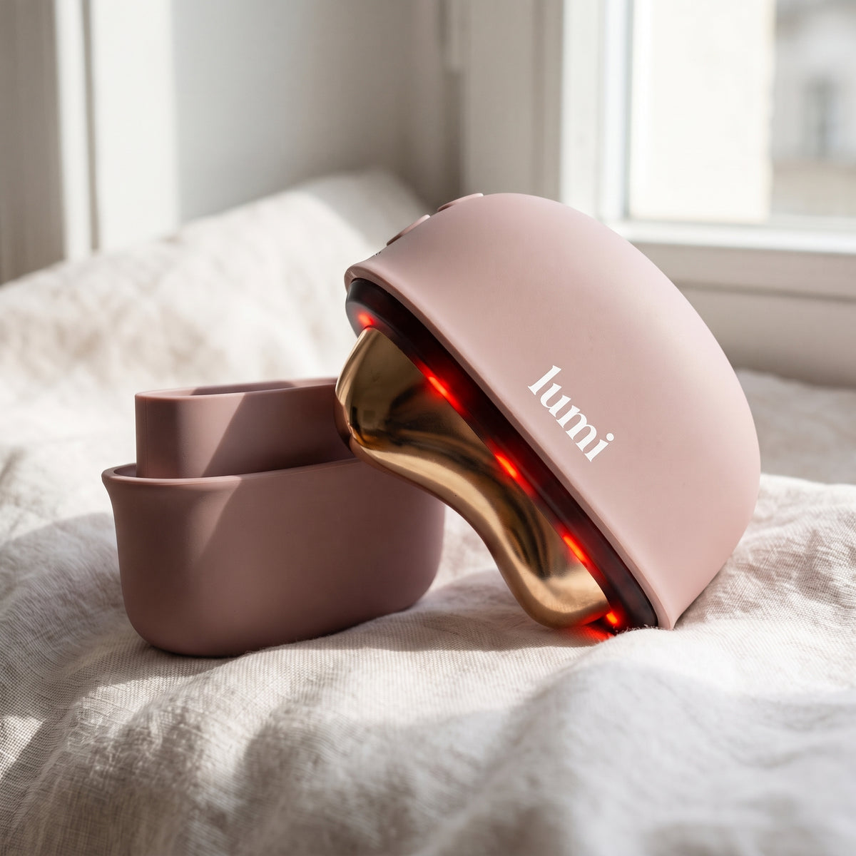 Lumi Ember rosé heated gua sha with its protective cover resting on soft linen by a window